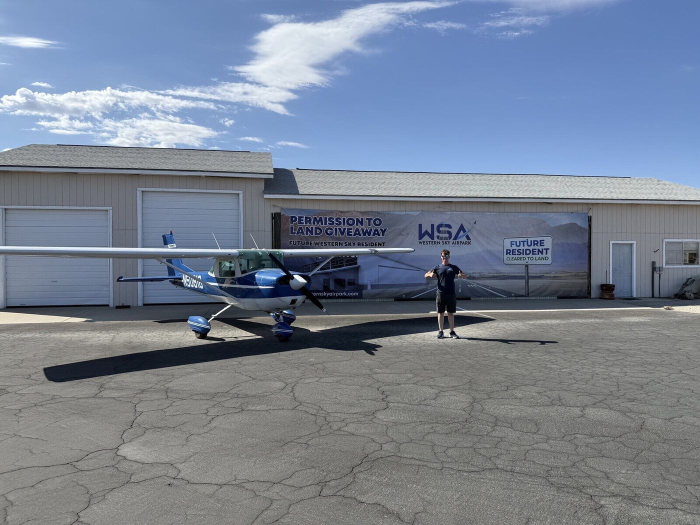 Hayden Bradley standing with his Cessna in front of the Permission to Land Giveaway banner at Western Sky Airpark